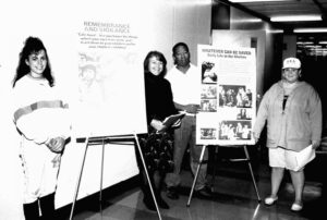 Three college-age students (two women who appear white and man who appears to be Black) and a teacher who appears to be a woman standing with two posters on easels. One reads, "Remembrance and Vigilance." The other reads, "Whatever Can be Saved: Daily Life in the Ghettos."