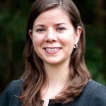 Headshot of Rebecca Root in a dark gray blazer. She is a white woman with long brown hair and small hoop earings.