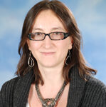 Headshot of Dr. Mia Serban against a blue background. She is wearing a dark grey sweater-blazer, a heavy necklace, and glasses. She is a white woman with medium-length brown hair. 