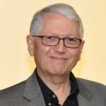 Headshot of Michael Riff wearing a black button-down shirt and a grey sports jacket. He is a middle aged white man with gray hair and glasses.