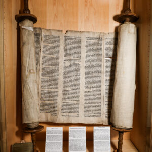 Picture of an opened Torah scroll in a wooden cabinet. There are three small informational plaques beneath it.