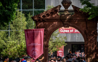 Students in caps and gowns walk down Mansion Road and under the Havemeyer Arch. Guests stand along either side to celebrate.