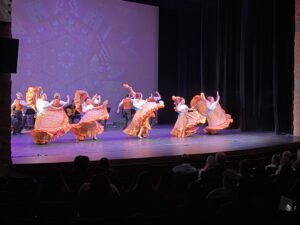 Five dancers in long, flowy skirts dance in a line across the stage