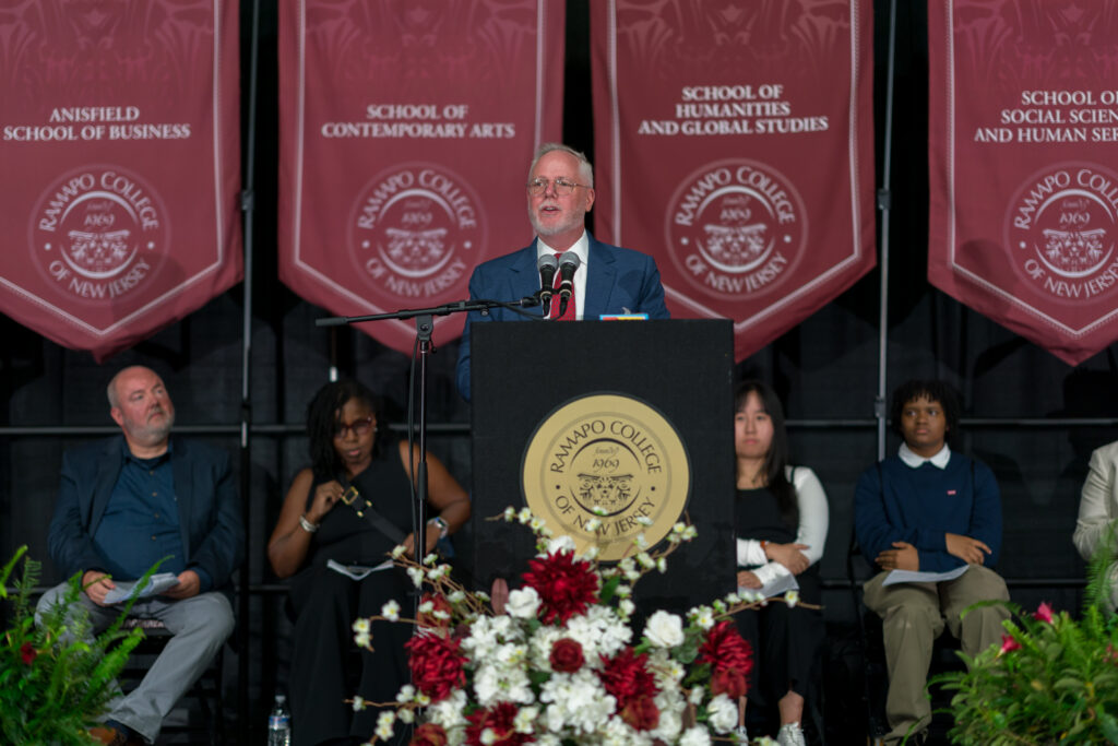 Provost Michael Middleton speaks to the Class of 2028 at the lectern on stage with maroon school banners hanging in the background. 