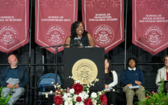 Author Victoria Christopher Murray speaks at the lectern addressing the Class of 2028. Maroon banners hang in the background on stage.