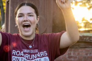 Student cheers in a Ramapo t-shirt in front of the Havemeyer Arch with the sun and trees in the background.