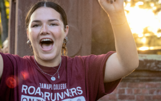 Student cheers in a Ramapo t-shirt in front of the Havemeyer Arch with the sun and trees in the background.