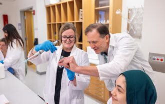 Two students work with test tubes in protective lab gear with guidance from their professor.