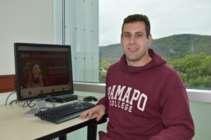 Wearing a Ramapo College hoodie, Christopher Barbieri '25 sits at a desk with a computer on it. A window displaying the Ramapo Mountains is behind him.