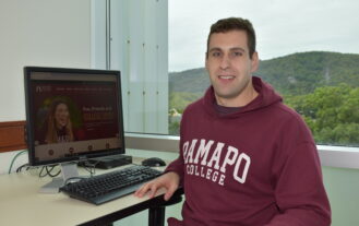 Wearing a Ramapo College hoodie, Christopher Barbieri '25 sits at a desk with a computer on it. A window displaying the Ramapo Mountains is behind him.