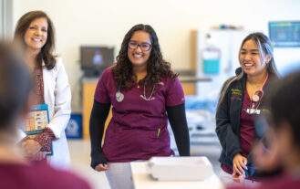 Two students in maroon and white scrubs with stethoscopes around their necks smile as they speak ot classmates in the simulation lab alongside a nursing faculty member who is wearing a white lab coat.