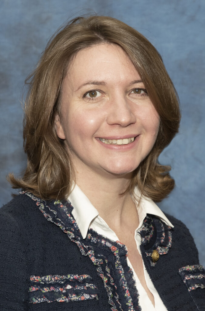 Image: Headshot of Dr. Natalia Santamaria-Laorden smiling at the camera in a blue, ruffled jacket and white, collared shirt with a blue background