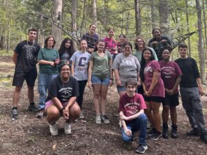 RCNJ students in the LIS program pose together in the woods in three rows with some kneeling in front, all standing in the middle, and some sitting in the rope net in the back.