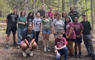 RCNJ students in the LIS program pose together in the woods in three rows with some kneeling in front, all standing in the middle, and some sitting in the rope net in the back.