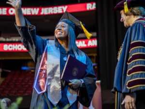 A female graduate stops to wave on the stage at Commencement after receiving her diploma cover. She waves to the crowd, which is behind us. The College President smiles and looks on from the right of the frame.