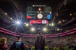 Image taken from the stage looking out over the arena floor filled with hundreds of graduates and seats filled with thousands of guests. The electronic banner on the big screen hanging from the ceiling reads "Congratulations Class of 2024!" President Jebb is standing at the podium. Her back is to the camera but she is facing the stage on the big screen.