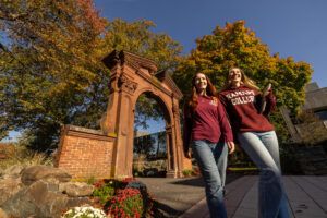 Two students wearing Ramapo College shirts walk off-camera with the Havemeyer Arch behind them.