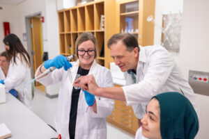 Two students work with test tubes in protective lab gear with guidance from their professor.