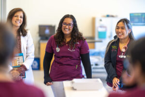 Two students in maroon and white scrubs with stethoscopes around their necks smile as they speak ot classmates in the simulation lab alongside a nursing faculty member who is wearing a white lab coat.