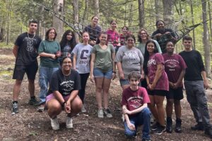 RCNJ students in the LIS program pose together in the woods in three rows with some kneeling in front, all standing in the middle, and some sitting in the rope net in the back.