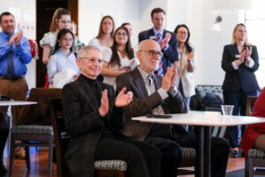 Susan and Nick Vallario are seated at a small table, clapping to someone off frame right. People behind them in the background are blurred and clapping.